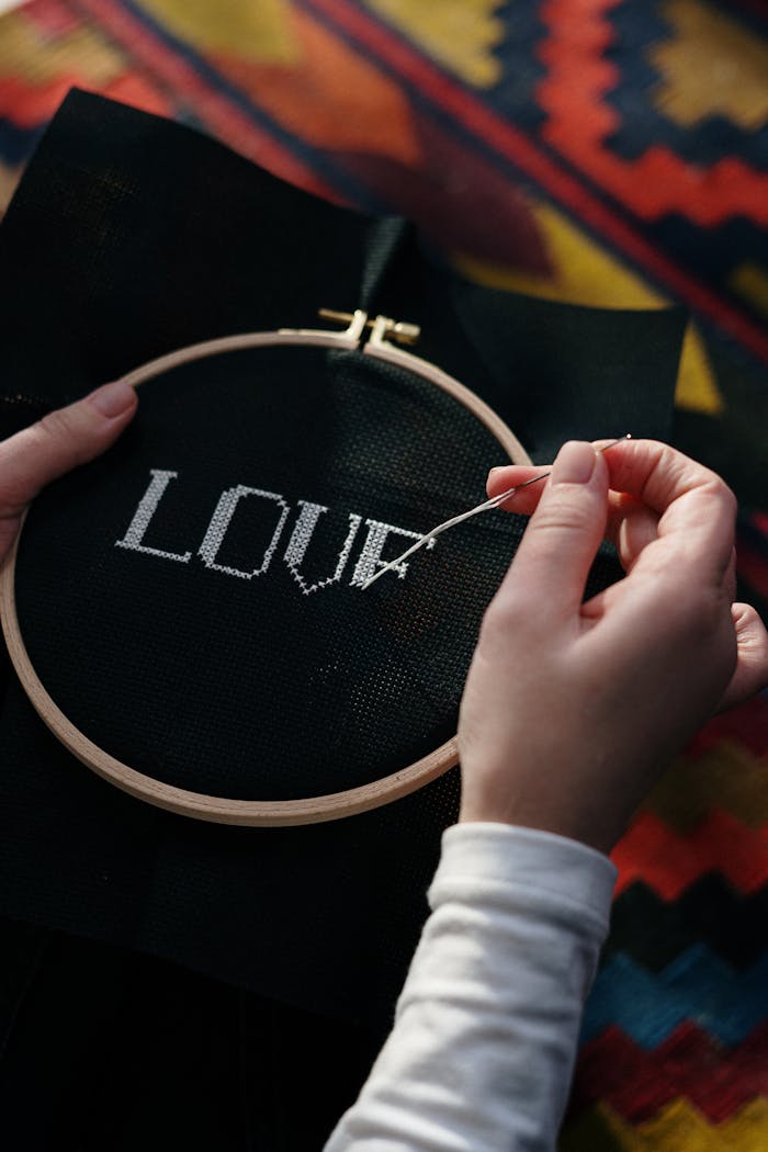 Close-up of hands embroidering 'LOVE' onto black fabric using a wooden hoop.